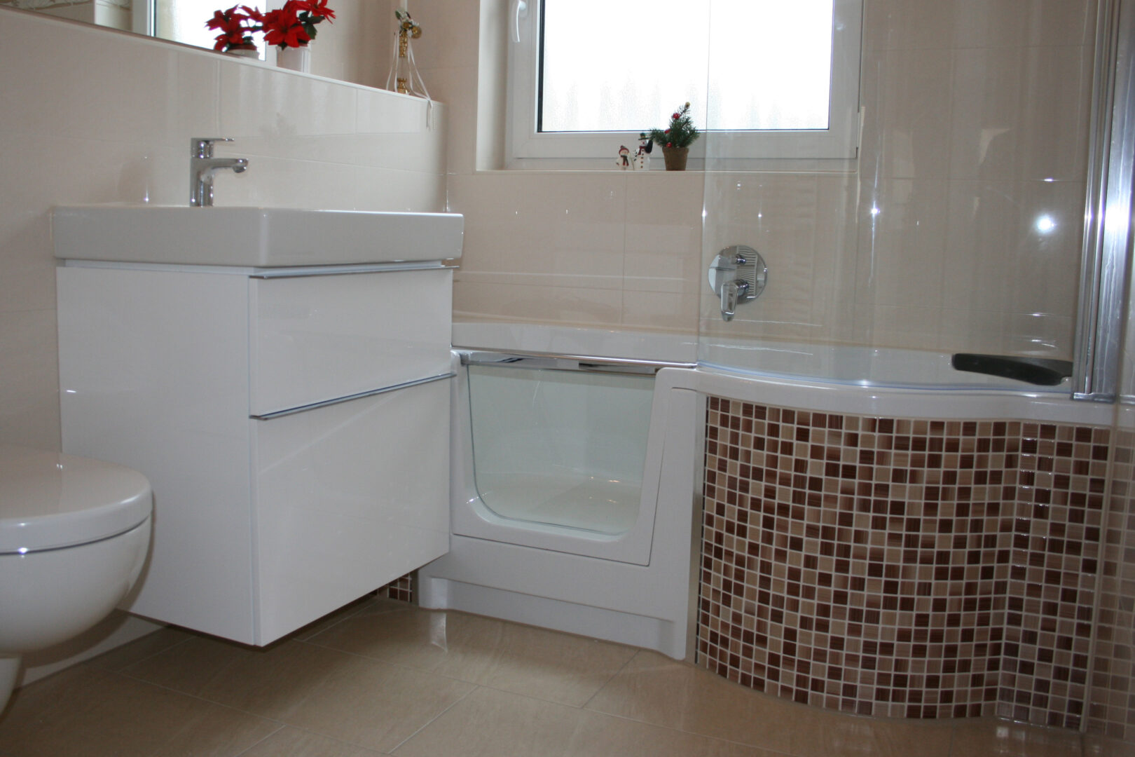 Barrierefreie Badewanne mit Einstiegstür und Mosaikfliesen, daneben Waschtisch und WC